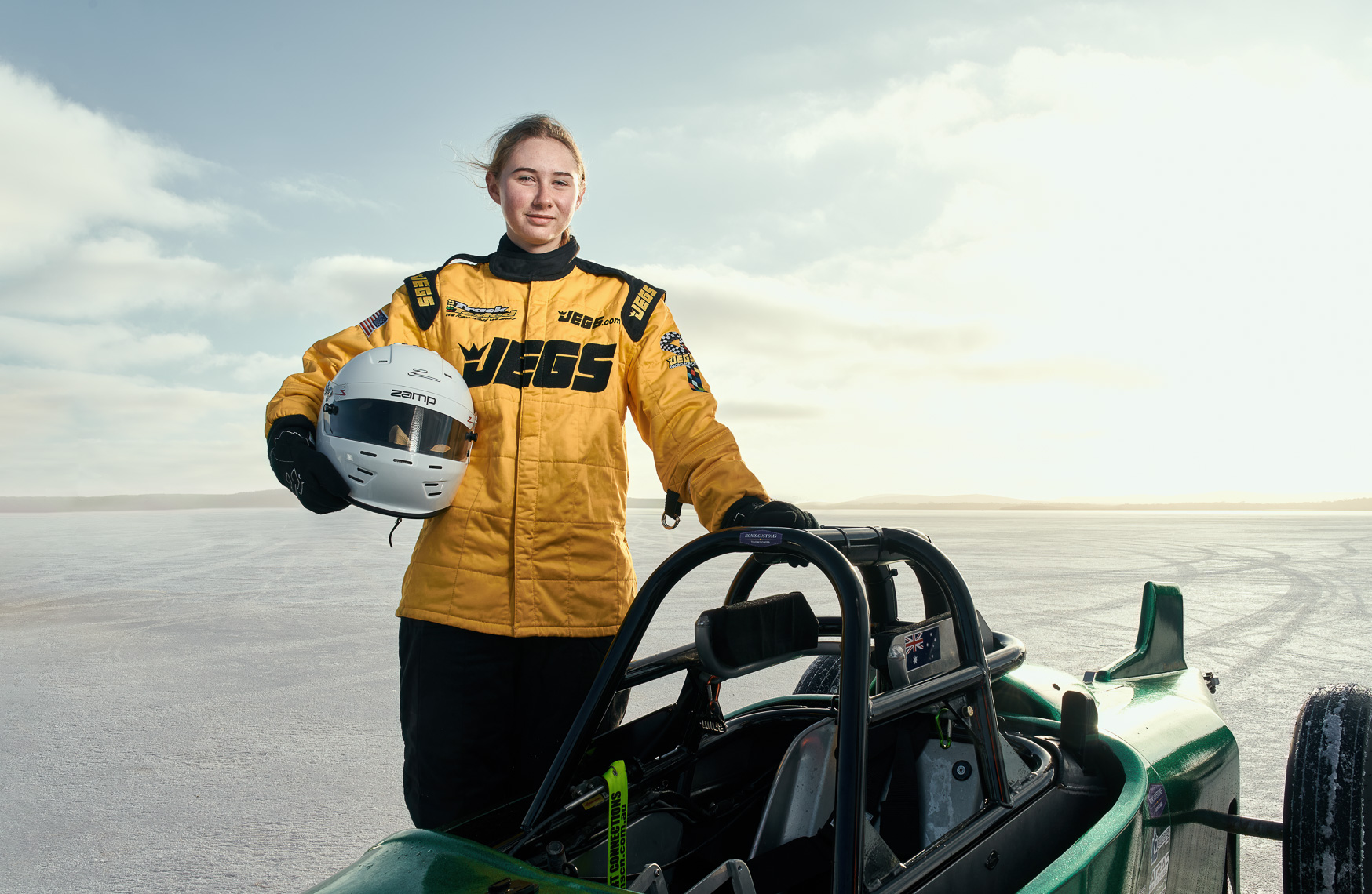 A teenage girl wearing a yellow race suit stands next to her racecar on a salt lake