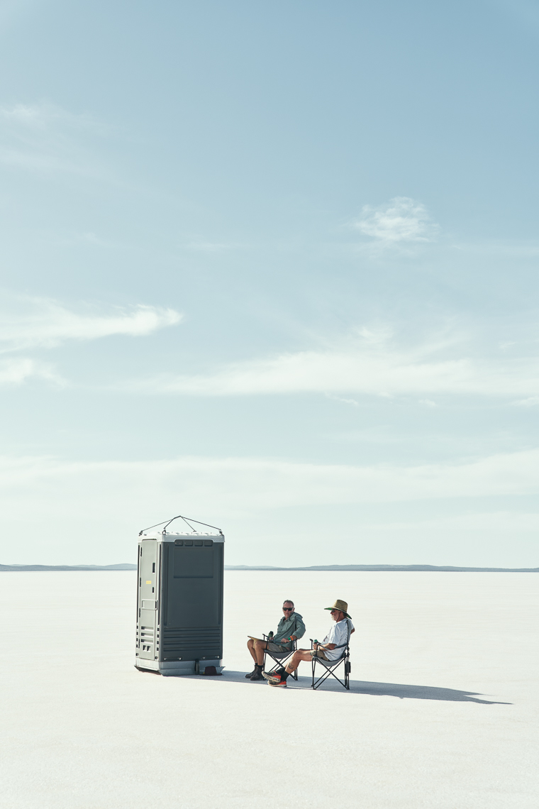 Two men sit in the shade of a portable toilet to hide from the harsh sun