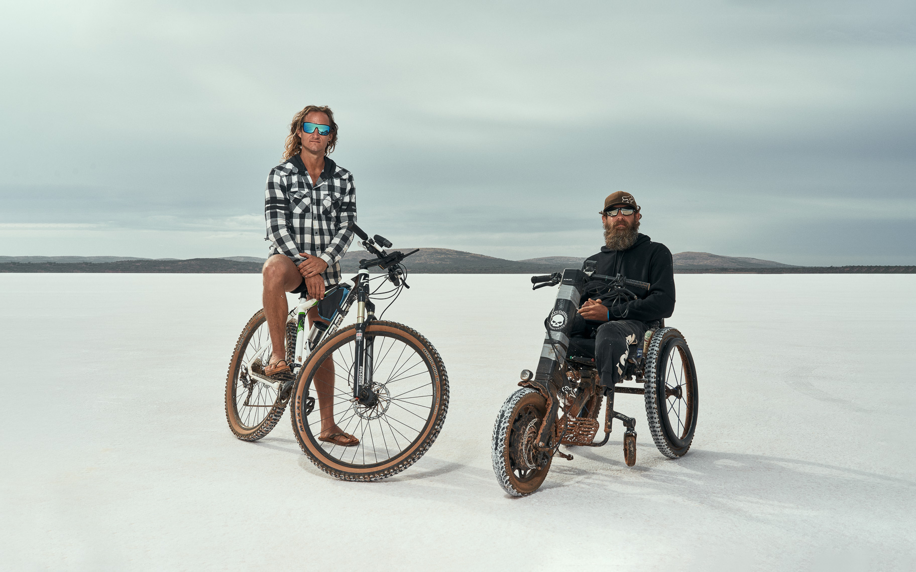 Two male friends sit on a bicycle and wheelchair on a salt lake